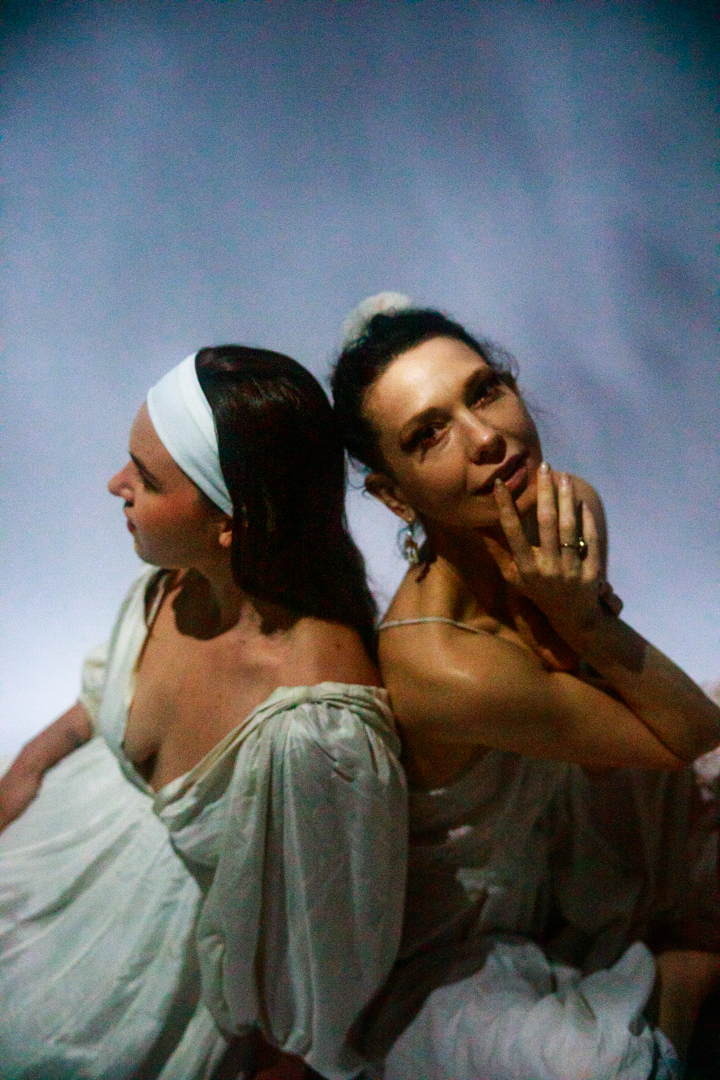 Two women sitting in the sky back to back, with one looking into the camera.